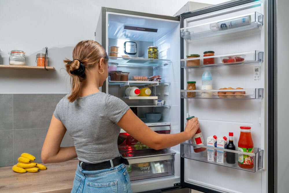 Osoba stoi przed otwartą lodówką, w której znajdują się różne produkty spożywcze, takie jak słoiki, butelki i jajka. Na blacie obok lodówki leżą banany, a na półce w tle stoją słoiki z przyprawami.