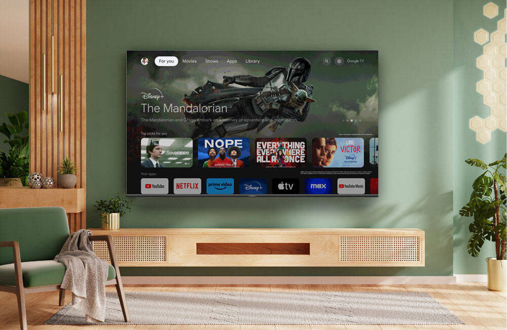 Telewizor wyświetla ekran główny z aplikacjami takimi jak YouTube, Netflix, Prime Video, Disney+, Apple TV, i max. Na ścianie wisi duży telewizor, a poniżej znajduje się drewniana szafka RTV.