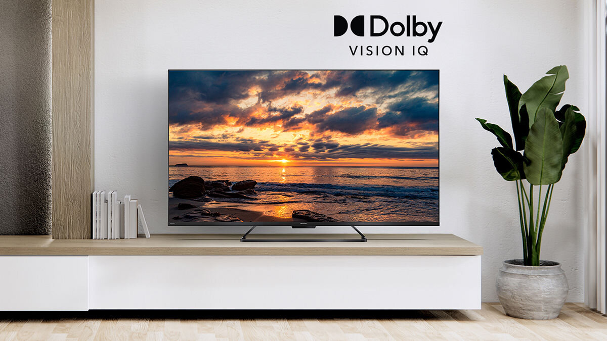 Telewizor wyświetla obraz zachodu słońca nad morzem, ustawiony na białej szafce. Na ścianie widoczne jest logo \'Dolby Vision IQ\'.