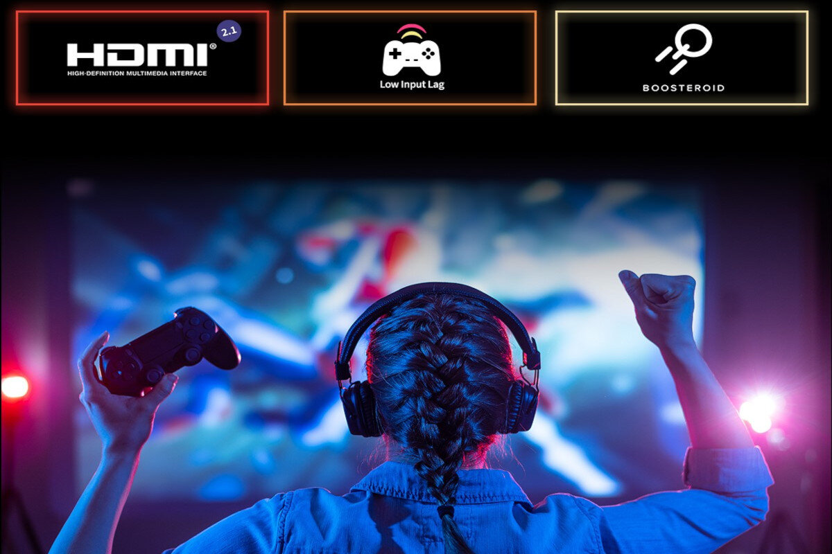 Osoba z kontrolerem do gier w rękach stoi przed dużym ekranem, na którym wyświetlany jest dynamiczny obraz. Na górze widoczne są trzy ikony z napisami: \'HDMI 2.1\', \'Low Input Lag\', \'BOOSTEROID\'.