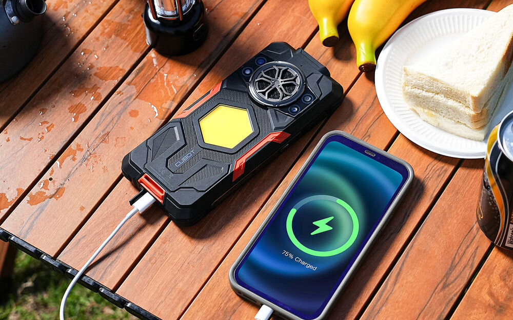 Na drewnianym stole leży czarny smartfon z żółtym sześciokątem na obudowie, podłączony do ładowania. Obok znajduje się inny telefon z wyświetlaczem pokazującym ikonę ładowania i napis \'75% Charged\'.