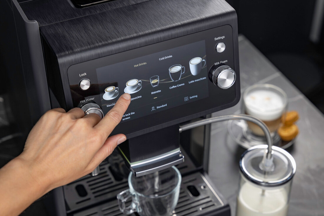 Osoba dotyka ekranu dotykowego ekspresu do kawy, na którym wyświetlane są opcje napojów takich jak \'Macchiato\', \'Espresso\', \'Coffee Creme\', \'Latte macchiato\'. Obok znajduje się pojemnik z mlekiem podłączony do ekspresu.