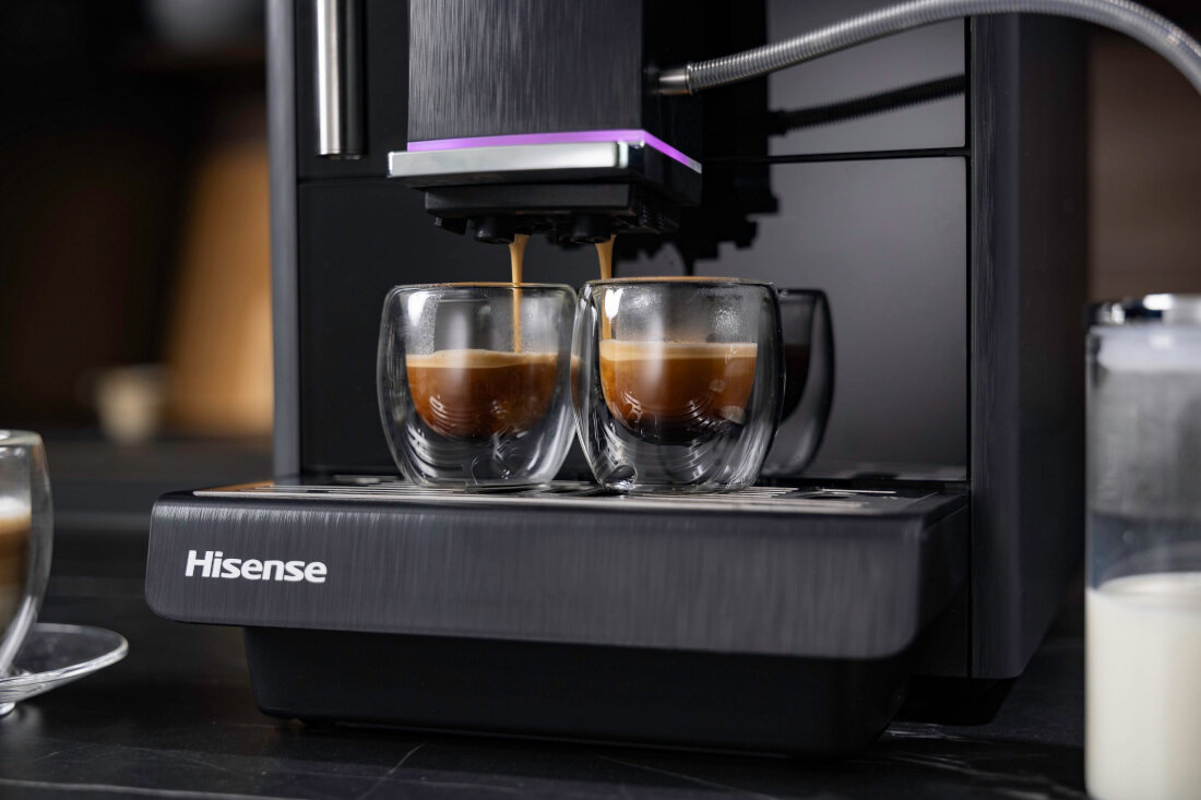 Ekspres do kawy z logo Hisense przygotowuje dwa espresso w szklankach. Obok znajduje się pojemnik z mlekiem.