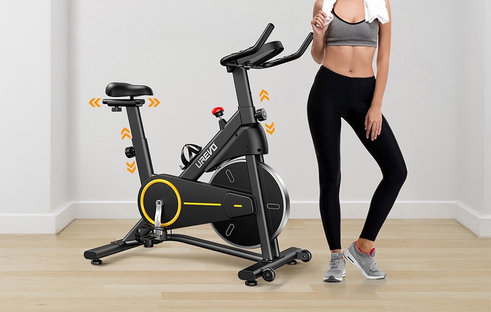 Na obrazie znajduje się czarny rower magnetyczny z napisem \'UREVO\' na ramie, ustawiony na drewnianej podłodze. Obok roweru stoi osoba w sportowym stroju, trzymająca ręcznik na ramionach.