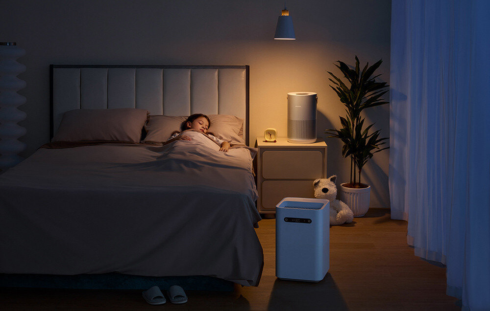 W sypialni znajduje się łóżko z osobą śpiącą, obok stoi nawilżacz powietrza na szafce nocnej. W tle widać roślinę doniczkową i pluszowego misia na podłodze.