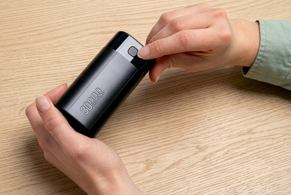 Osoba trzyma czarny powerbank z napisem \'30000 mAh\' na drewnianym stole. Na urządzeniu widoczny jest przycisk z symbolem włączania.