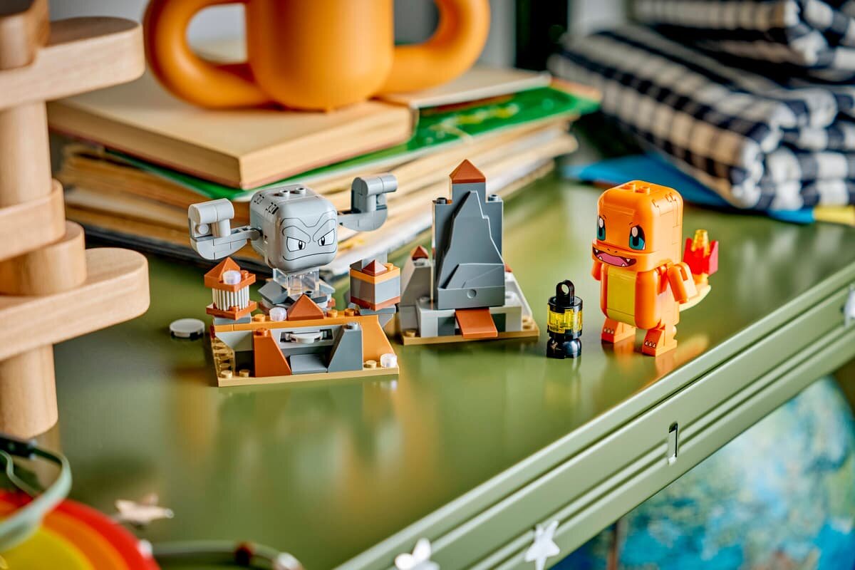 Na zielonym stole znajdują się klocki LEGO przedstawiające postacie i elementy krajobrazu, w tym pomarańczowy stworek z ognistym ogonem oraz szary, kamienny stwór. W tle widoczne są książki i inne przedmioty domowe.