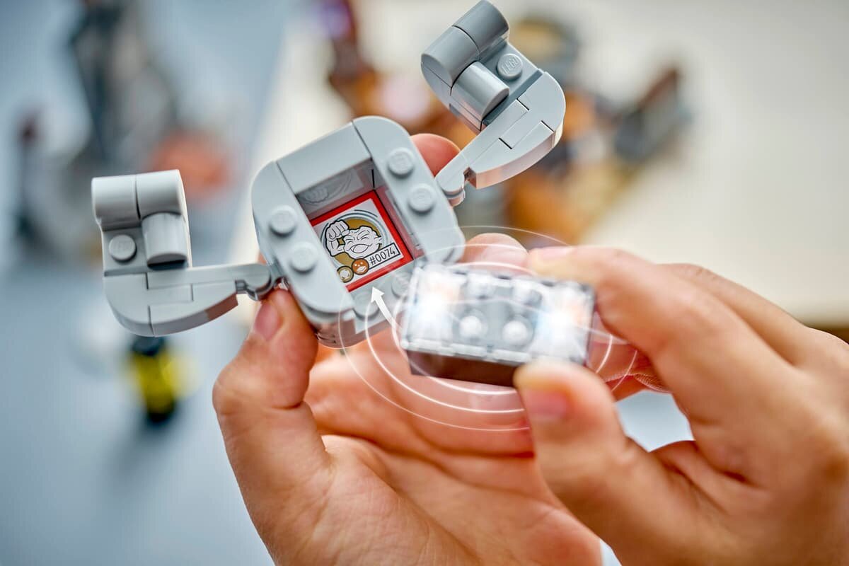 Osoba trzyma szary element konstrukcyjny z klocków LEGO, na którym widoczny jest rysunek postaci z napisem \'0074\'.