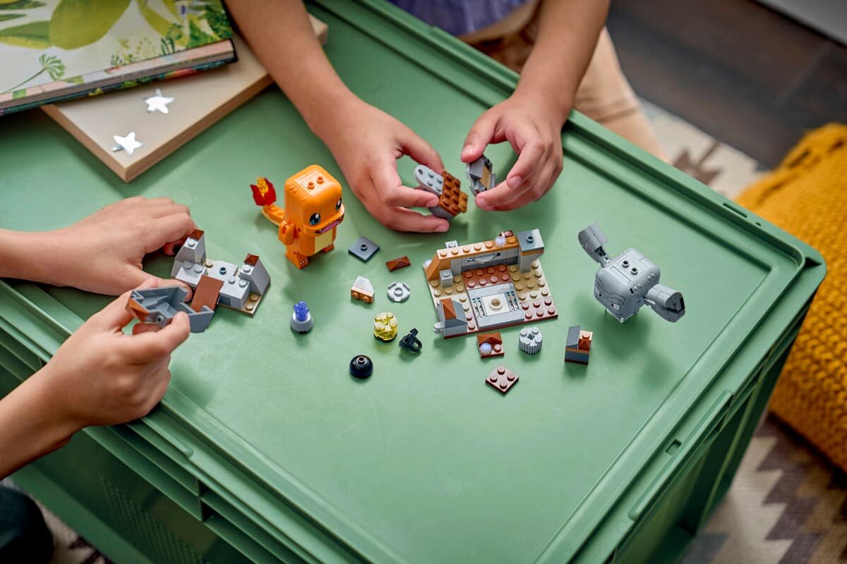 Na zielonym stole znajdują się klocki LEGO, w tym figurki przypominające postacie z gry Pokémon. Dwie osoby bawią się zestawem, układając elementy konstrukcyjne.