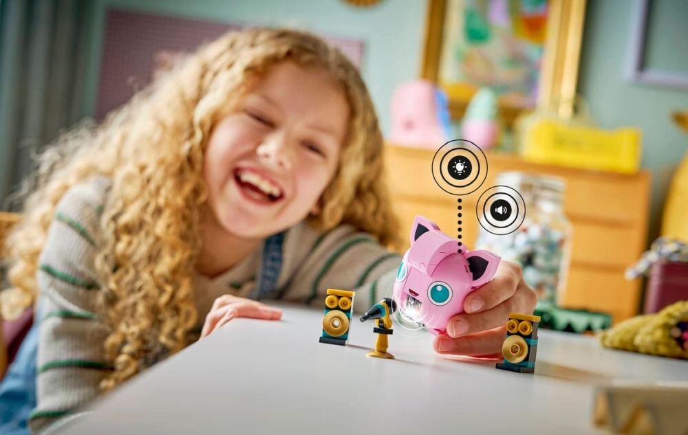 Dziecko bawi się różowym modelem Jigglypuff z serii LEGO, otoczonym przez małe głośniki i mikrofon. W tle widać rozmyte elementy pokoju dziecięcego.