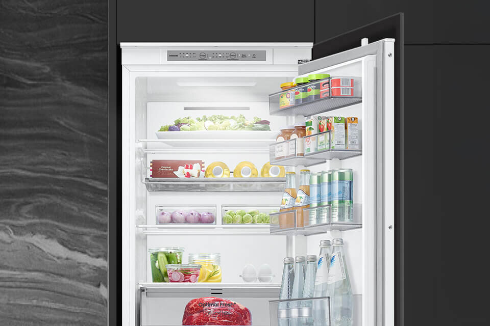 Otwarta lodówka z widocznymi półkami wypełnionymi różnorodnymi produktami spożywczymi, takimi jak warzywa, napoje i jajka. Na drzwiach lodówki znajdują się dodatkowe półki z kartonami i butelkami.