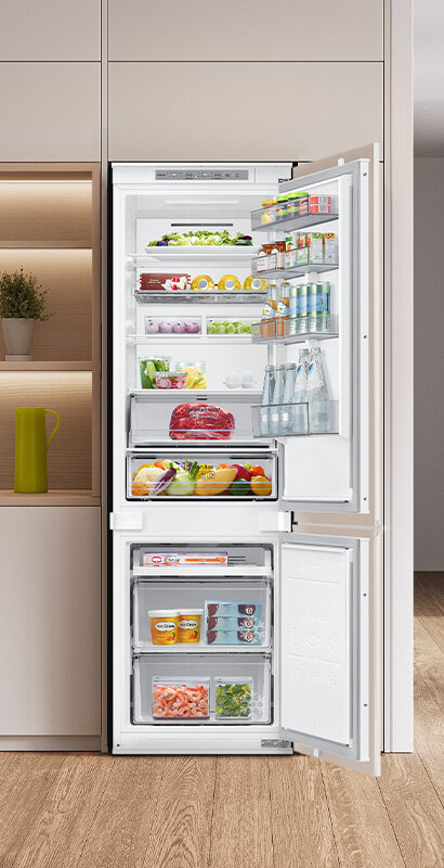 Otwarta lodówka z widocznymi półkami wypełnionymi różnorodnymi produktami spożywczymi, takimi jak owoce, warzywa, napoje i mięso. Wnętrze jest jasne i dobrze zorganizowane, z szufladami i półkami na drzwiach.