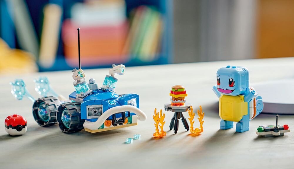 Na stole znajdują się klocki LEGO przedstawiające niebieskiego stworka z dużymi oczami, pojazd terenowy z czterema kołami oraz mały model ogniska z płomieniami. Obok widoczna jest kulka przypominająca Poké Ball oraz mały kontroler.
