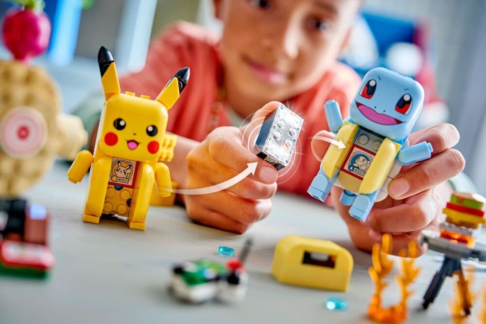 Na obrazie widać dwie figurki LEGO przedstawiające postacie z Pokémon: Pikachu i Squirtle. W tle znajduje się dziecko, a na stole rozłożone są różne elementy LEGO, w tym ogień i jedzenie.