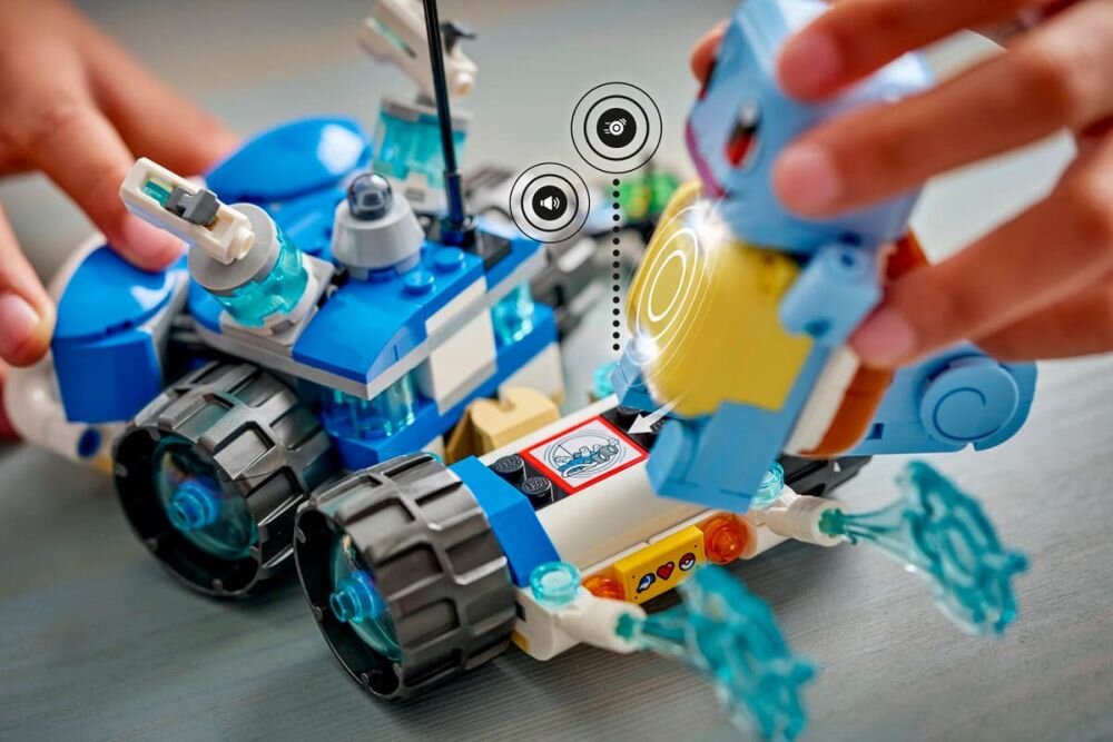 Na obrazie widać zestaw klocków LEGO przedstawiający niebieski pojazd terenowy z dużymi kołami i figurką przypominającą postać z gry Pokémon. Obok pojazdu znajdują się ikony symbolizujące funkcje interaktywne.