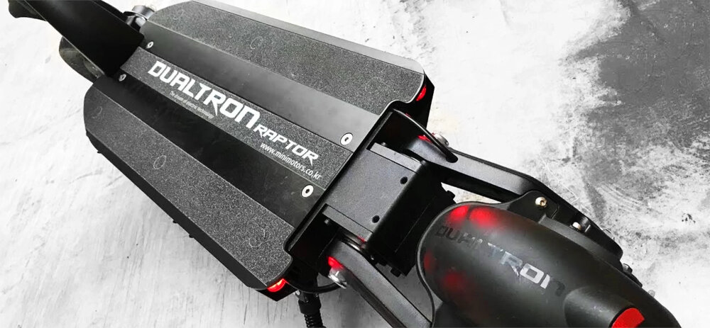 Czarna hulajnoga elektryczna z widocznym napisem \'DUALTRON Raptor\' na platformie. W tle widać szarą powierzchnię.
