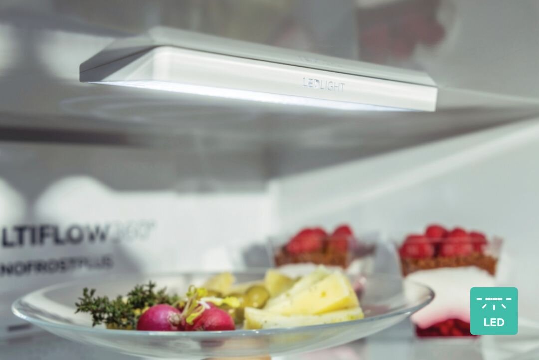 Wnętrze lodówki z oświetleniem LED, na półce znajduje się talerz z warzywami i ziołami. Widoczny jest napis \'LED LIGHT\' oraz \'MULTIFLOW 360° NOFROST PLUS\'.