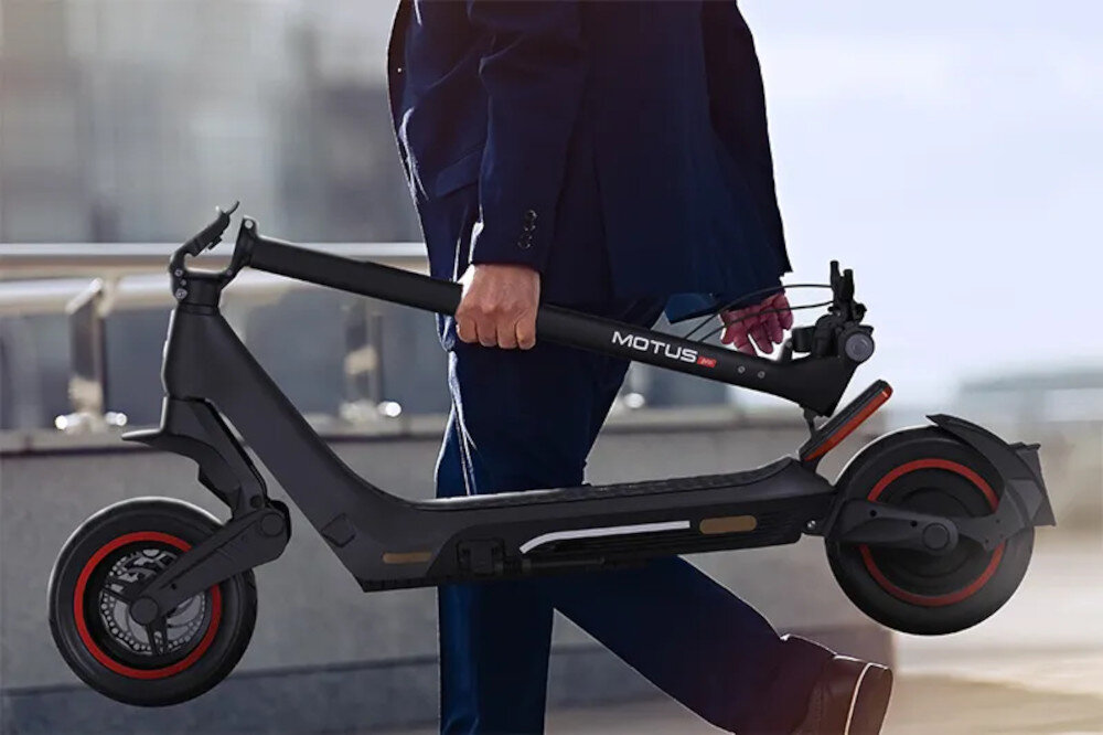 Osoba w garniturze niesie złożoną hulajnogę elektryczną z widocznym napisem \'MOTUS\'. Hulajnoga ma czarne wykończenie z czerwonymi akcentami na kołach.