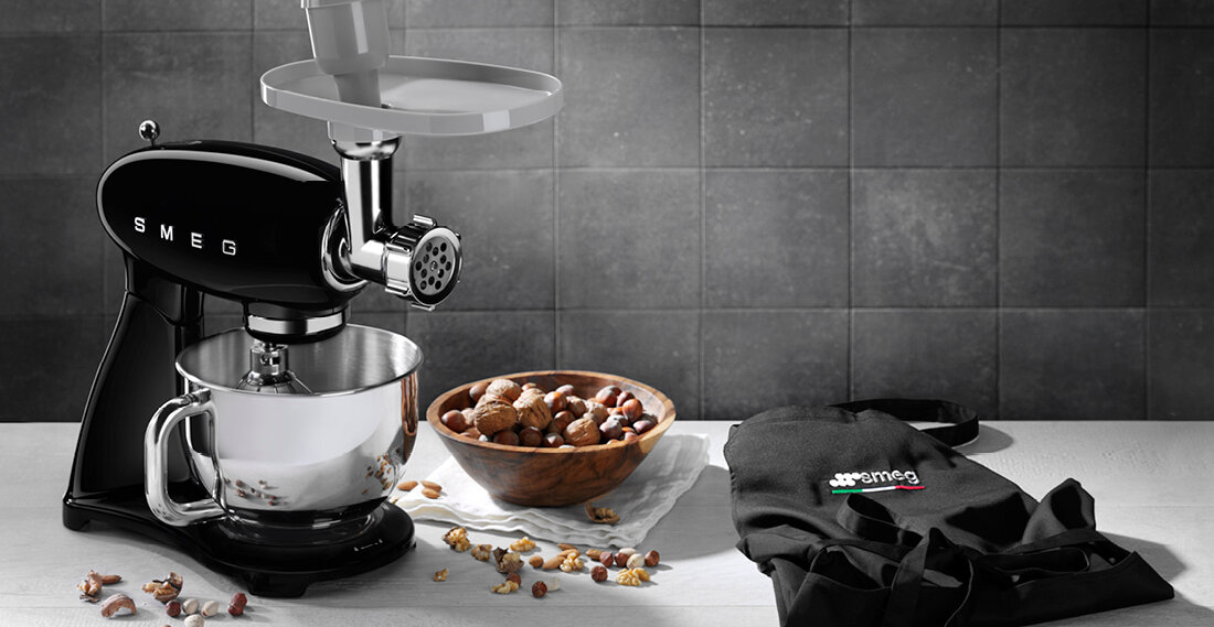 Na blacie kuchennym stoi czarny robot kuchenny z logo SMEG, obok znajduje się miska z orzechami oraz czarna torba z logo SMEG. W tle widoczna jest szara, kafelkowa ściana.