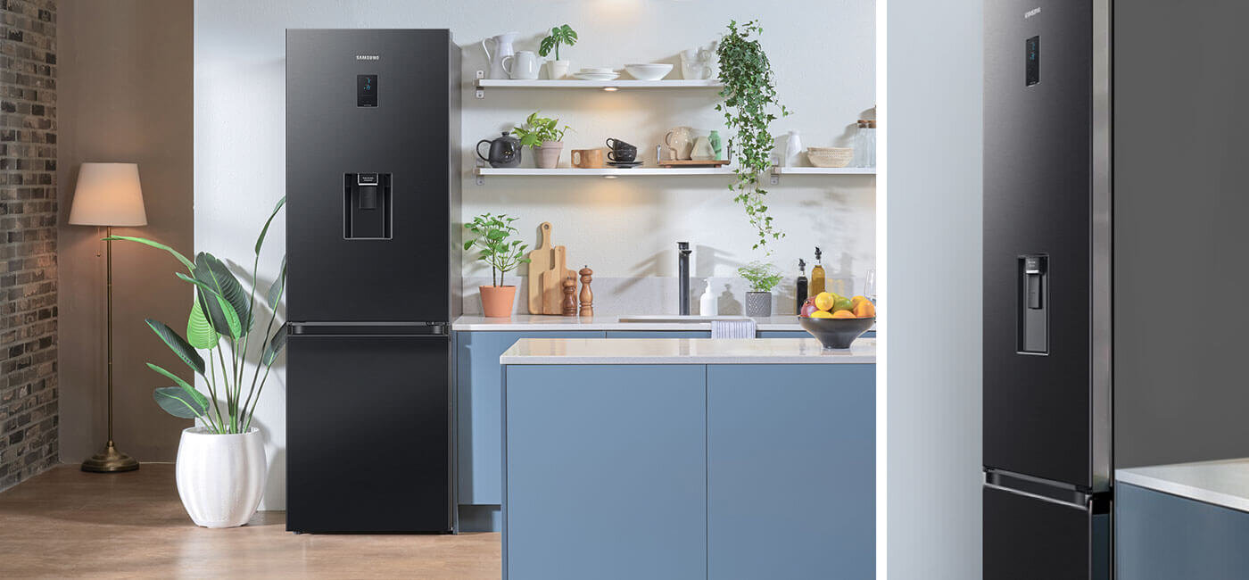 Design lodówki RB34C652EBN dopasowany do nowoczesnej kuchni Czarna lodówka z podajnikiem wody stoi w nowoczesnej kuchni z niebieskimi szafkami i roślinami na półkach. Na lodówce widoczne jest logo Samsung.