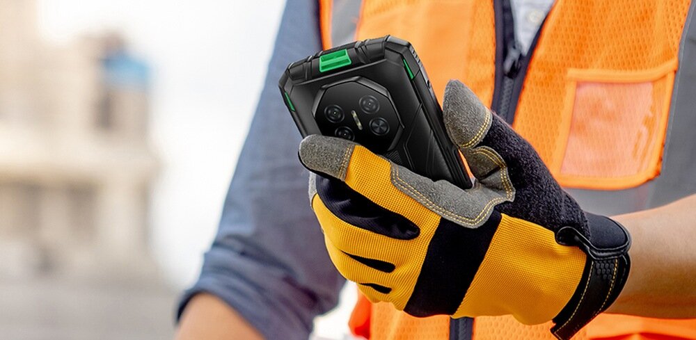 Osoba w pomarańczowej kamizelce i rękawicach ochronnych trzyma smartfon z czarną obudową i zielonymi akcentami. Telefon ma widoczne cztery obiektywy aparatu na tylnej części.