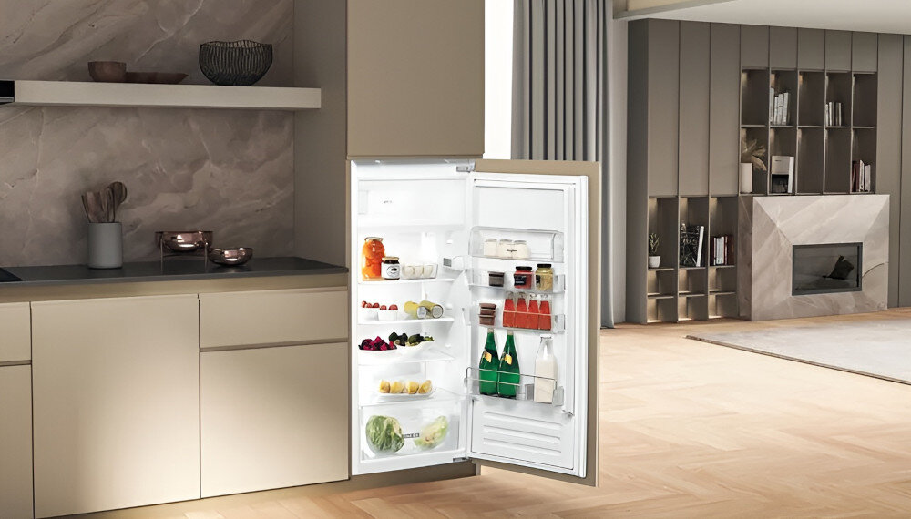 Wnętrze nowoczesnej kuchni z otwartą lodówką, w której znajdują się różne produkty spożywcze, takie jak napoje, owoce i słoiki. W tle widoczny jest salon z kominkiem i półkami na książki.