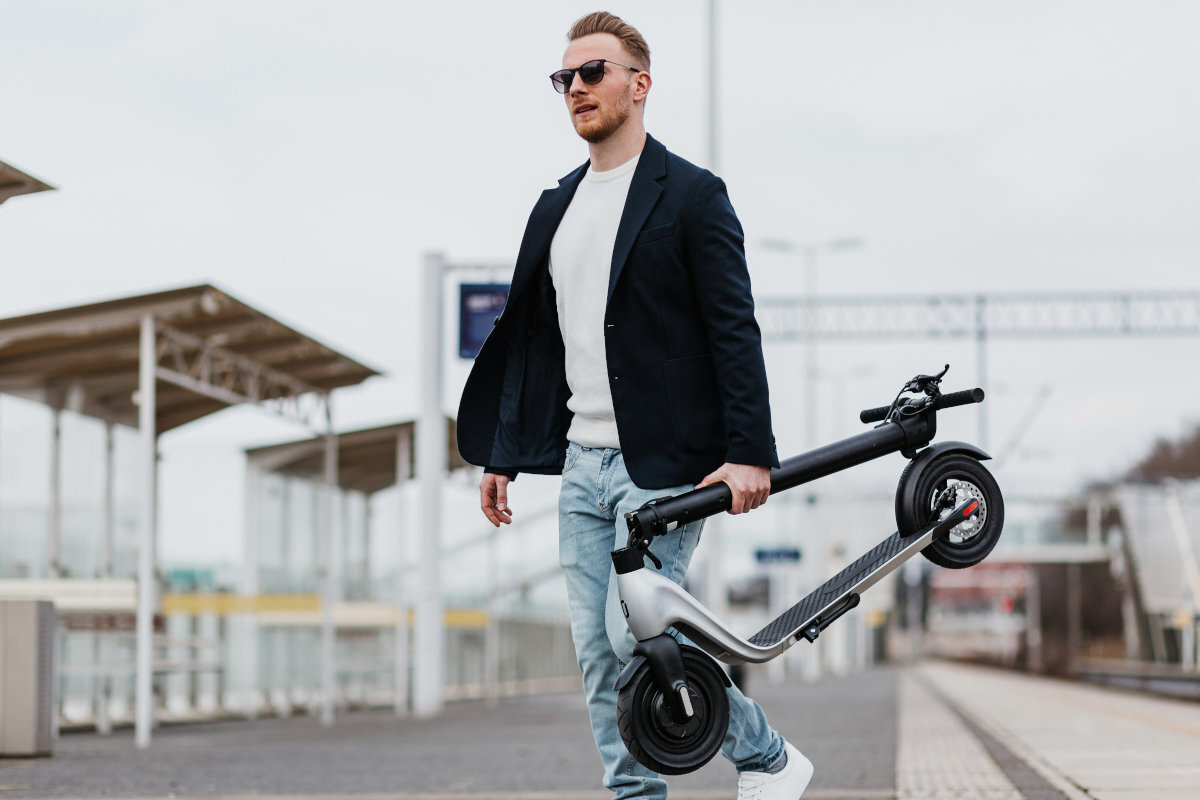 Mężczyzna w okularach przeciwsłonecznych niesie złożoną hulajnogę elektryczną w kolorze czarno-srebrnym na peronie kolejowym. W tle widoczne są zadaszenia i tory kolejowe.