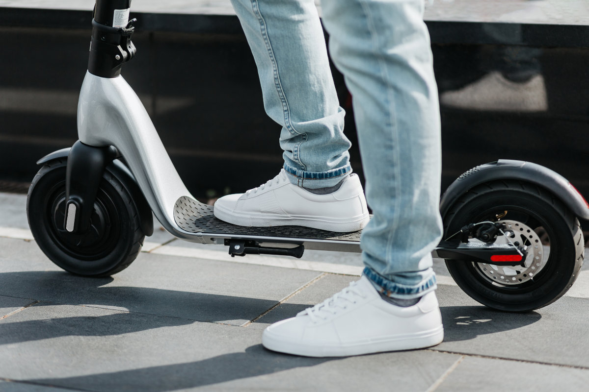 Osoba stoi na elektrycznej hulajnodze o czarno-srebrnym kolorze, ubrana w jasne jeansy i białe buty. Hulajnoga ma duże koła i widoczny jest jej mechanizm hamulcowy.