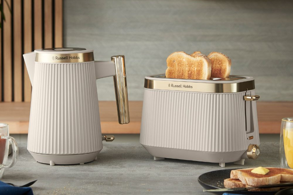 Na obrazie widoczny jest czajnik i toster marki Russell Hobbs w jasnym kolorze z metalicznymi elementami. Toster zawiera dwie kromki chleba, a obok znajduje się szklanka z sokiem pomarańczowym i talerz z tostami.