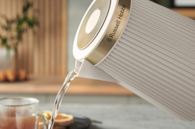 Biały czajnik z logo \'Russell Hobbs\' nalewa wodę do szklanki. W tle widoczne są rozmyte elementy kuchenne.