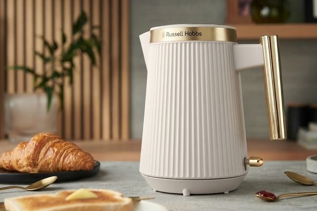 Na stole znajduje się elegancki czajnik z pionowymi żłobieniami i złotymi elementami, z widocznym logo \'Russell Hobbs\'. Obok leży talerz z rogalikiem oraz kromka chleba z masłem.