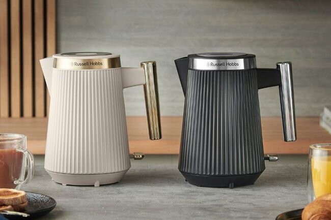 Na blacie stoją dwa czajniki elektryczne z logo \'Russell Hobbs\', jeden w kolorze beżowym, drugi czarnym. Obok znajdują się filiżanka z herbatą i szklanka z sokiem pomarańczowym.