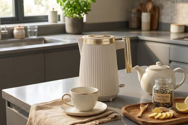 Na blacie kuchennym znajduje się biały czajnik elektryczny z logo \'Russell Hobbs\', obok filiżanka na spodku oraz dzbanek do herbaty. Na drewnianej tacy leży słoik z napisem \'Silver Needle White Tea\' oraz plasterki imbiru.