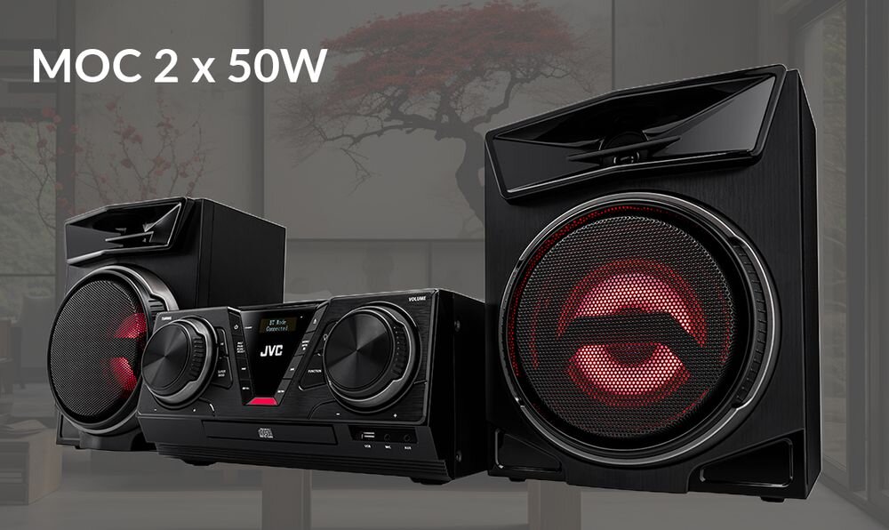 Wieża audio JVC z dwoma głośnikami, wyświetlaczem i pokrętłami, na tle wnętrza z drzewem za oknem. Widoczny tekst: \'MOC 2 x 50W\'.