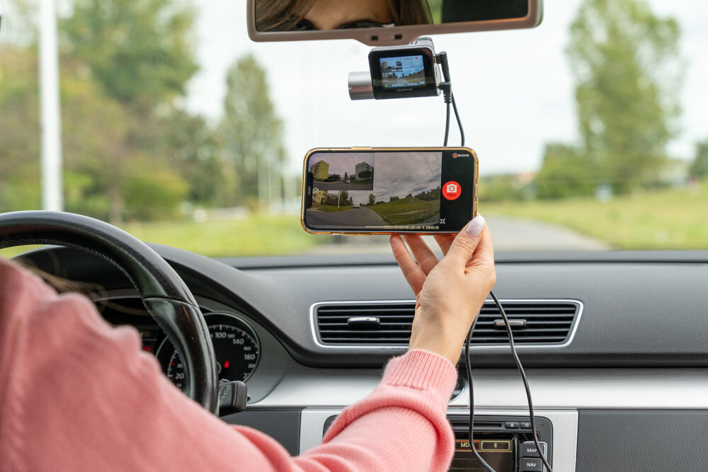 Osoba siedząca w samochodzie trzyma smartfon z włączoną aplikacją do nagrywania wideo, a na przedniej szybie zamontowany jest wideorejestrator. W tle widać drogę i zieleń za oknem samochodu.