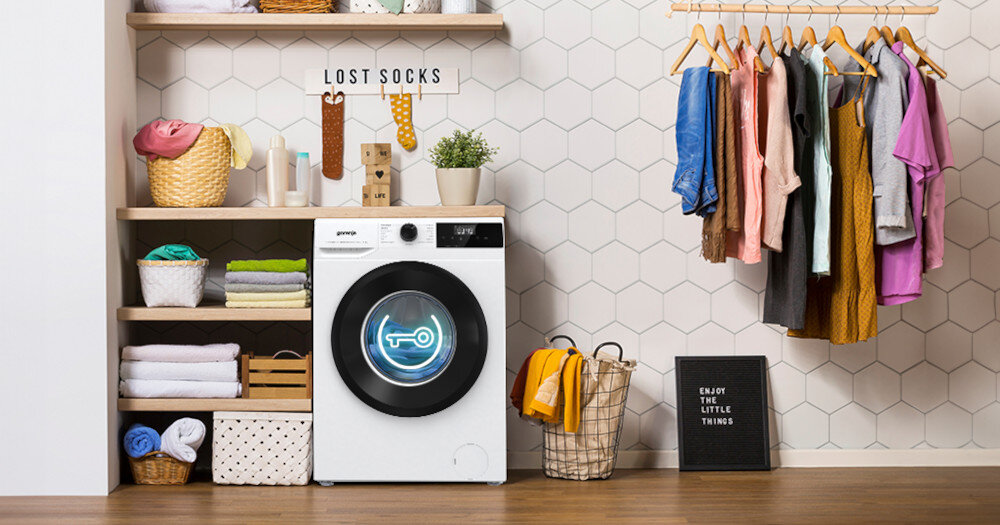 Pralka z widocznym logo producenta Gorenje stoi w pomieszczeniu z półkami pełnymi koszy i ręczników oraz wieszakami z ubraniami. Na ścianie znajduje się napis \'LOST SOCKS\', a obok pralki tabliczka z napisem \'ENJOY THE LITTLE THINGS\'.