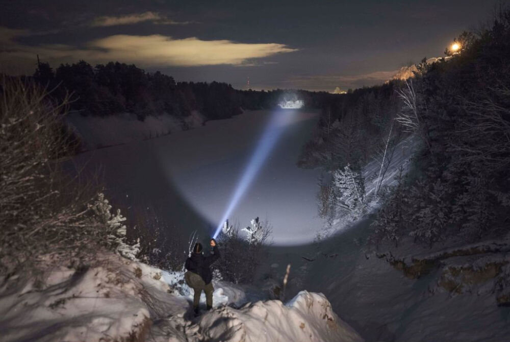 Osoba stoi na zaśnieżonym zboczu, oświetlając latarką zamarzniętą rzekę lub jezioro, otoczone drzewami. W tle widać ciemne niebo z chmurami.