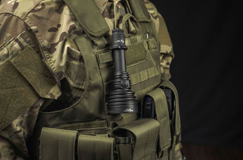Osoba w kamuflażu wojskowym nosi na kamizelce taktycznej czarną latarkę z napisem \'ARMYTEK\'.
