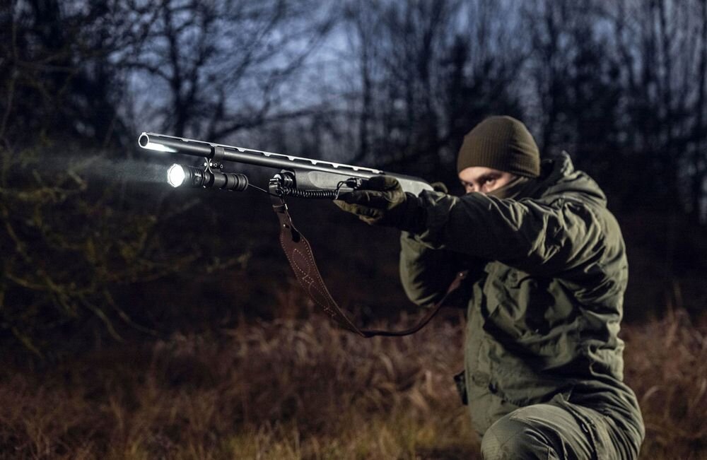 Latarka taktyczna ARMYTEK F08201PC Predator Pro Max Czarny, montaż, pokrowiec, klips, smycz, Mężczyzna celuje z broni z zamontowaną latarką.