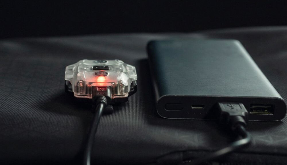 Na obrazie widać przezroczystą latarkę z czerwonym światłem podłączoną do czarnego powerbanku za pomocą kabla USB.