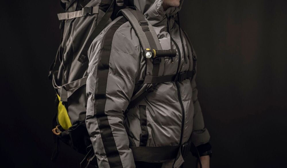 Latarka ARMYTEK F10702PC Elf C2 Max LR Czarny, wymiary, waga, magnes, klips, Mężczyzna w szarej kurtce z dużym plecakiem turystycznym, do którego szelki przymocowana jest czarna latarka kątowa.
