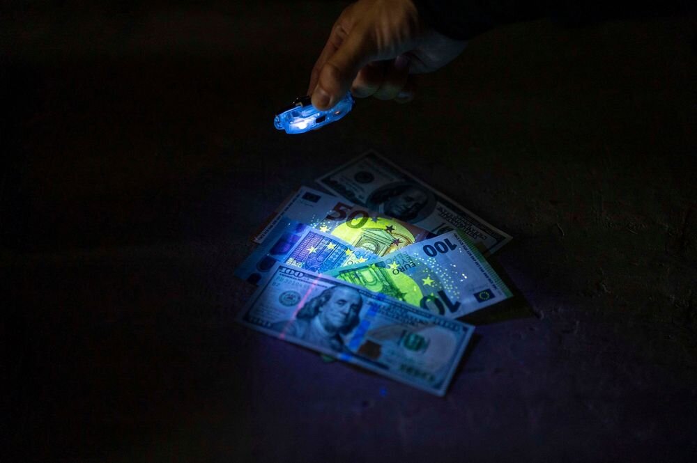 Ręka trzymająca latarkę UV oświetla banknoty dolarowe i euro, ukazując ich zabezpieczenia. Banknoty leżą na ciemnym tle.