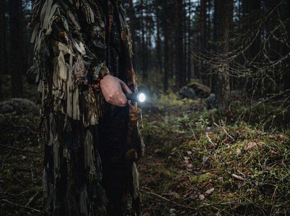 Latarka taktyczna ARMYTEK F02005C, filtry, soczewka, reflektor, optyka, Osoba w kamuflażu idzie przez las z włączoną latarką.
