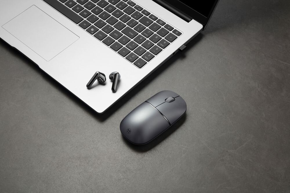 Na ciemnym blacie znajduje się otwarty laptop, obok którego leżą szare słuchawki bezprzewodowe i mysz komputerowa. Mysz ma logo producenta na powierzchni.