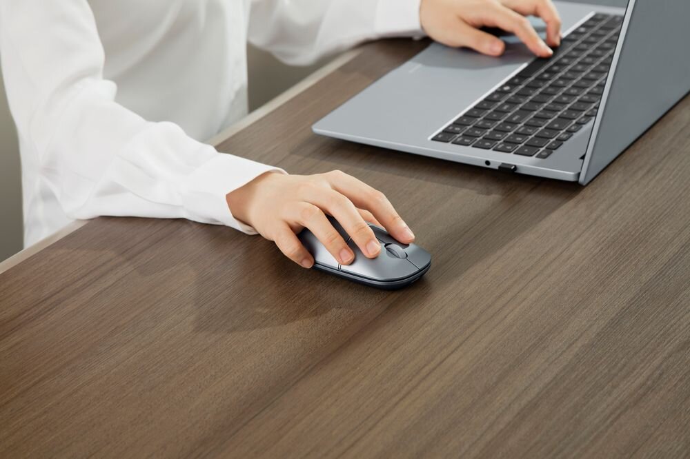 Osoba używa szarej myszy komputerowej przy laptopie na drewnianym biurku. Laptop jest otwarty, a osoba ma na sobie białą koszulę.