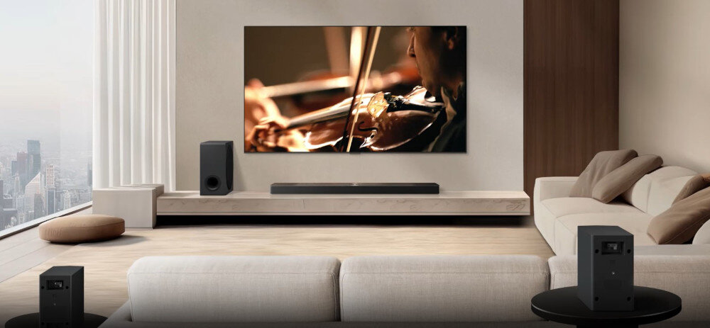 W nowoczesnym salonie znajduje się duży telewizor wyświetlający obraz skrzypiec, a pod nim umieszczony jest soundbar. W pomieszczeniu widoczne są także głośniki i jasne meble wypoczynkowe.