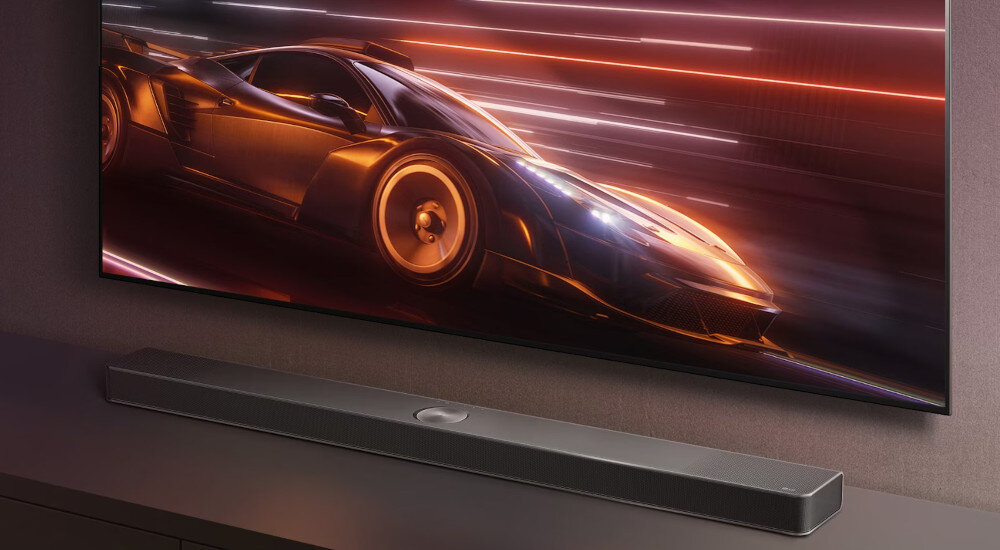 Na obrazie widoczny jest czarny soundbar umieszczony pod telewizorem, na którym wyświetlany jest dynamiczny obraz samochodu sportowego w ruchu. Soundbar ma prosty, nowoczesny design.