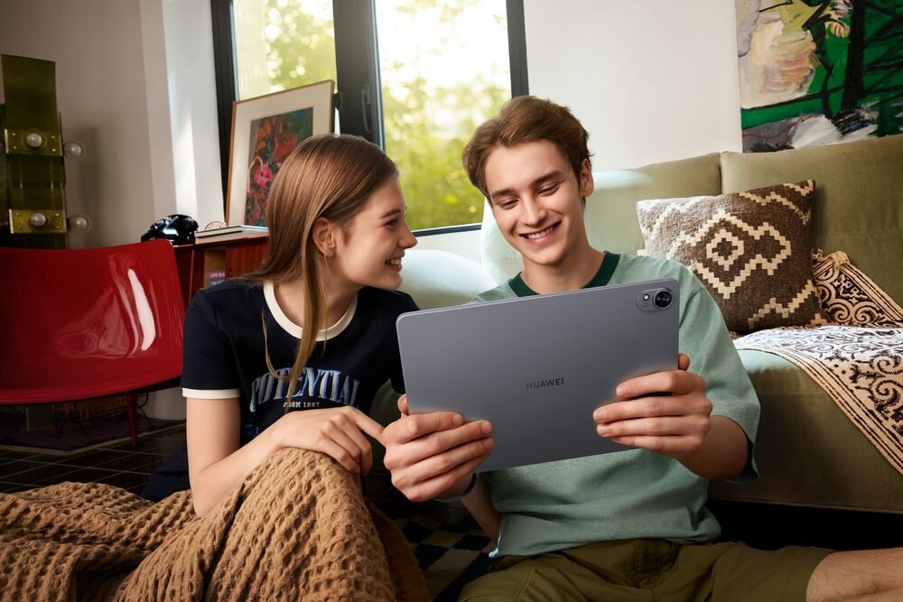 Dwie osoby siedzą na podłodze, trzymając tablet marki HUAWEI. W tle widać sofę, poduszki i okno z widokiem na zewnątrz.