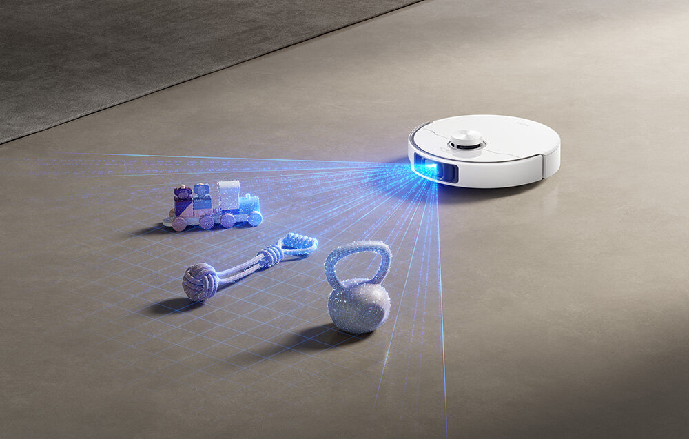 Na podłodze znajduje się biały robot sprzątający, który emituje niebieskie światło skanujące przedmioty, takie jak zabawka pociąg, sznur i kettlebell.