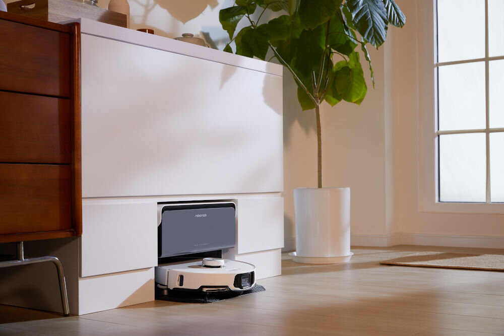 Robot sprzątający znajduje się w stacji dokującej pod białą szafką. Obok stoi duża roślina w białej donicy, a w tle widać okno.